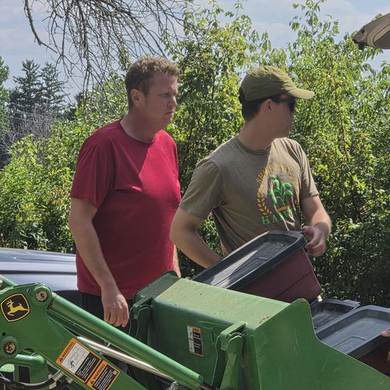 Two helpers unloading bins of collected ingredients into a tractor bucket